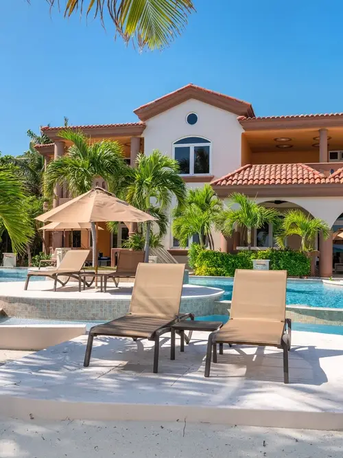 Pool deck with lounge chairs and umbrellas in front of a two-story Spanish-style building with a red tile roof